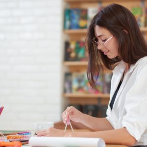 Charming young woman smiling and looking down. Female painter looking down and sitting at table with painter's supplies. Beautiful girl wearing in spectacles and white shirt.